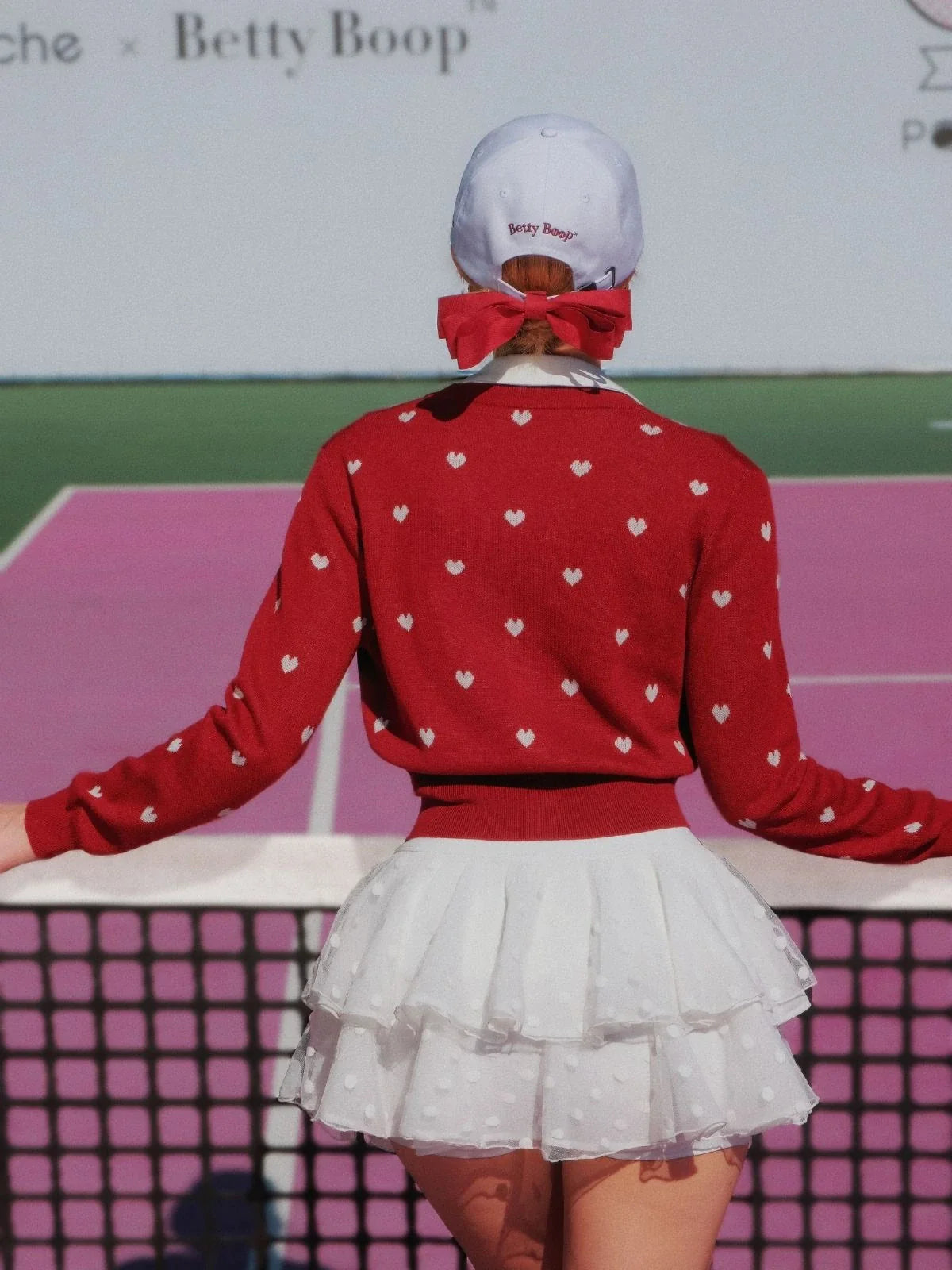 Person wearing a red sweater with white heart patterns and a white skirt on a tennis court.