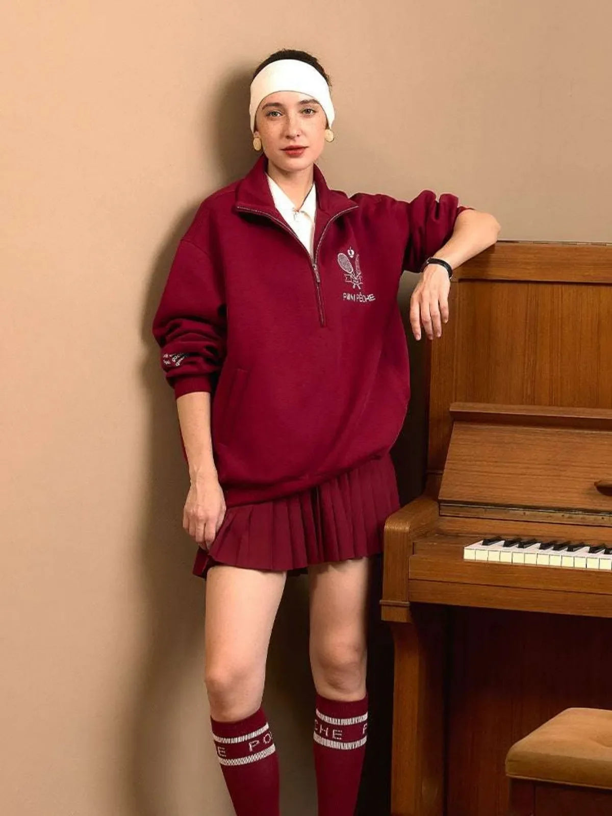 Person wearing a red outfit with a headband, standing next to a wooden piano against a beige wall.
