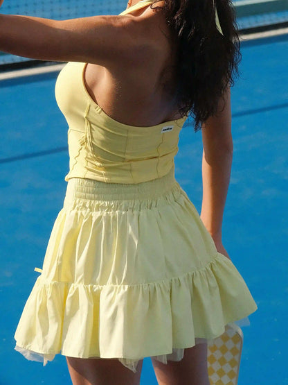 Yellow backless tennis top with a fitted silhouette and subtle logo detail, styled on the tennis court.
