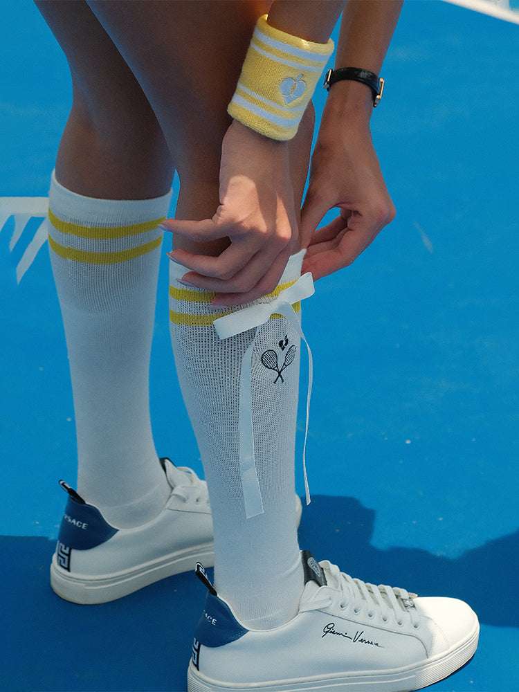 White knee-high tennis socks with light yellow stripes, bow detail, and logo embroidery.